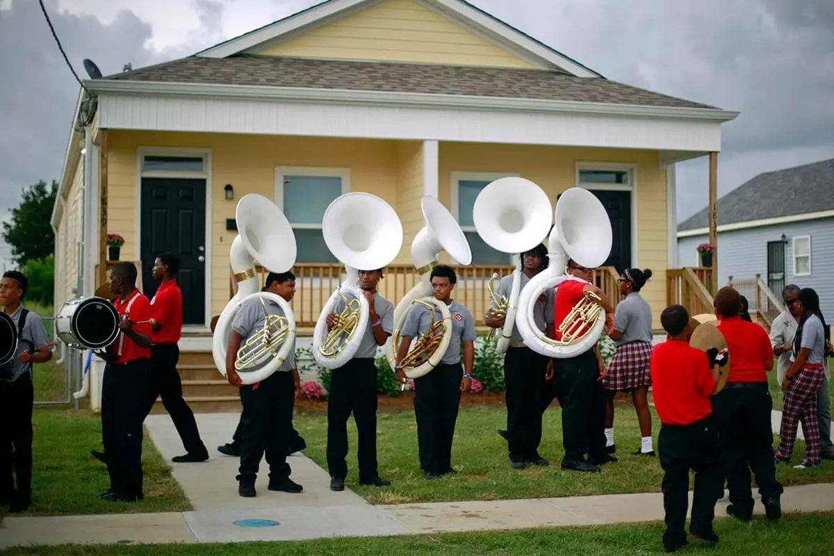 A marching band wearing red and black plays music in front of a yellow house